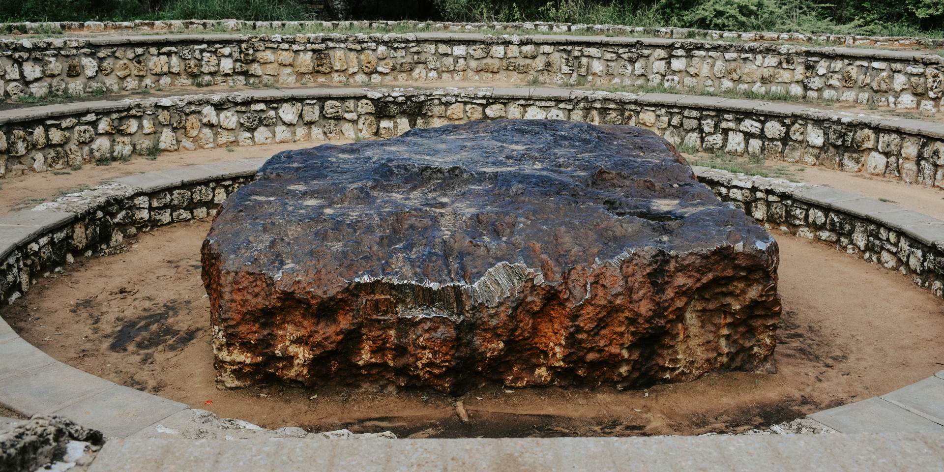 A massive iron meteorite partially buried in the ground, surrounded by open Namibian landscape and an amphitheatre for visitors.